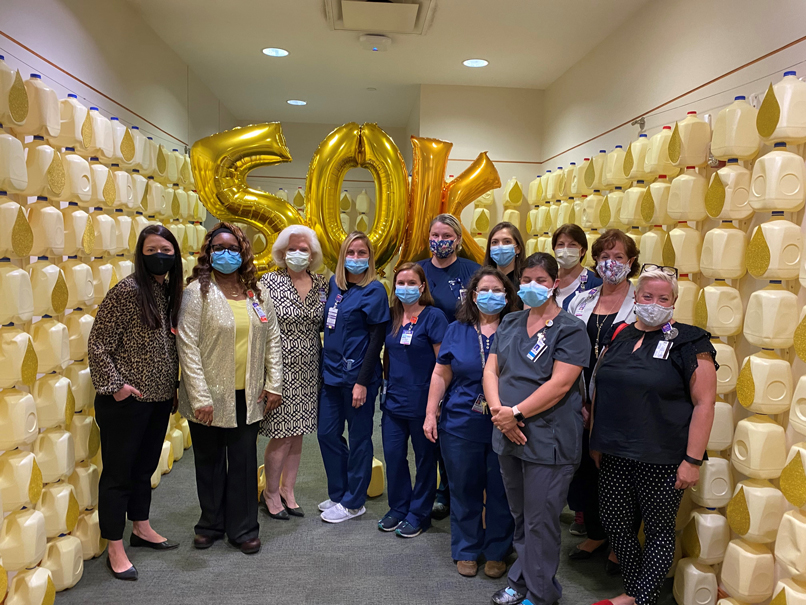 Team members wearing masks at milk jug display — containers on wall represent 50,000 gallons of donations. "50K" balloons in background.