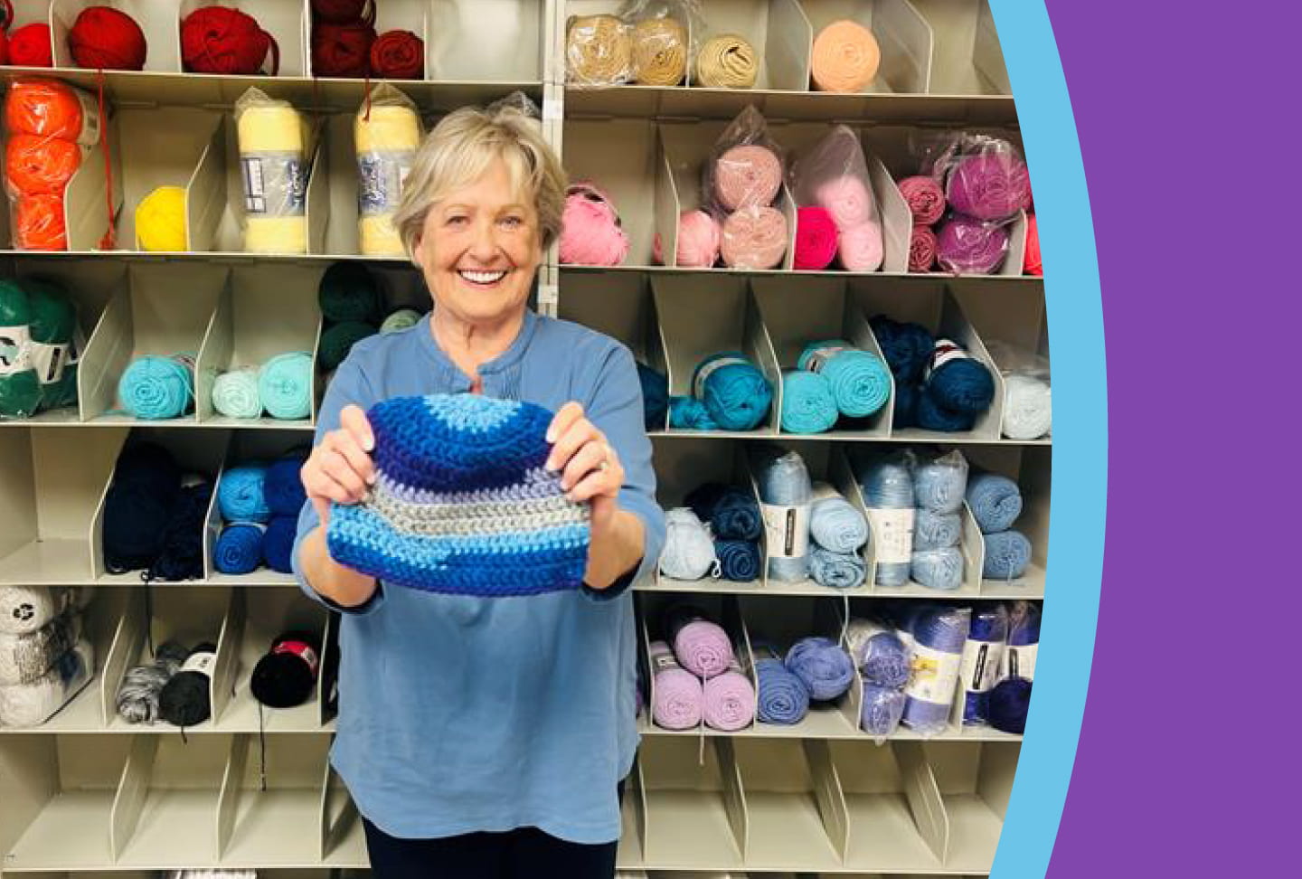 Kathy DeJoseph holding a knitted hat in front of wall of yarn