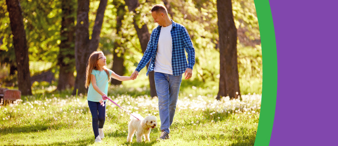 A father and daughter walk with their dog on a bright spring day.