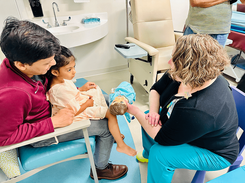 Shows a Child Life Specialist sharing information about a medical procedure for a child to understand, using a doll to demonstrate.