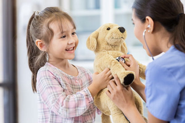 Shows a medical professional demonstrating how to use a stethoscope to a young child, using a teddy bear to demonstrate. 